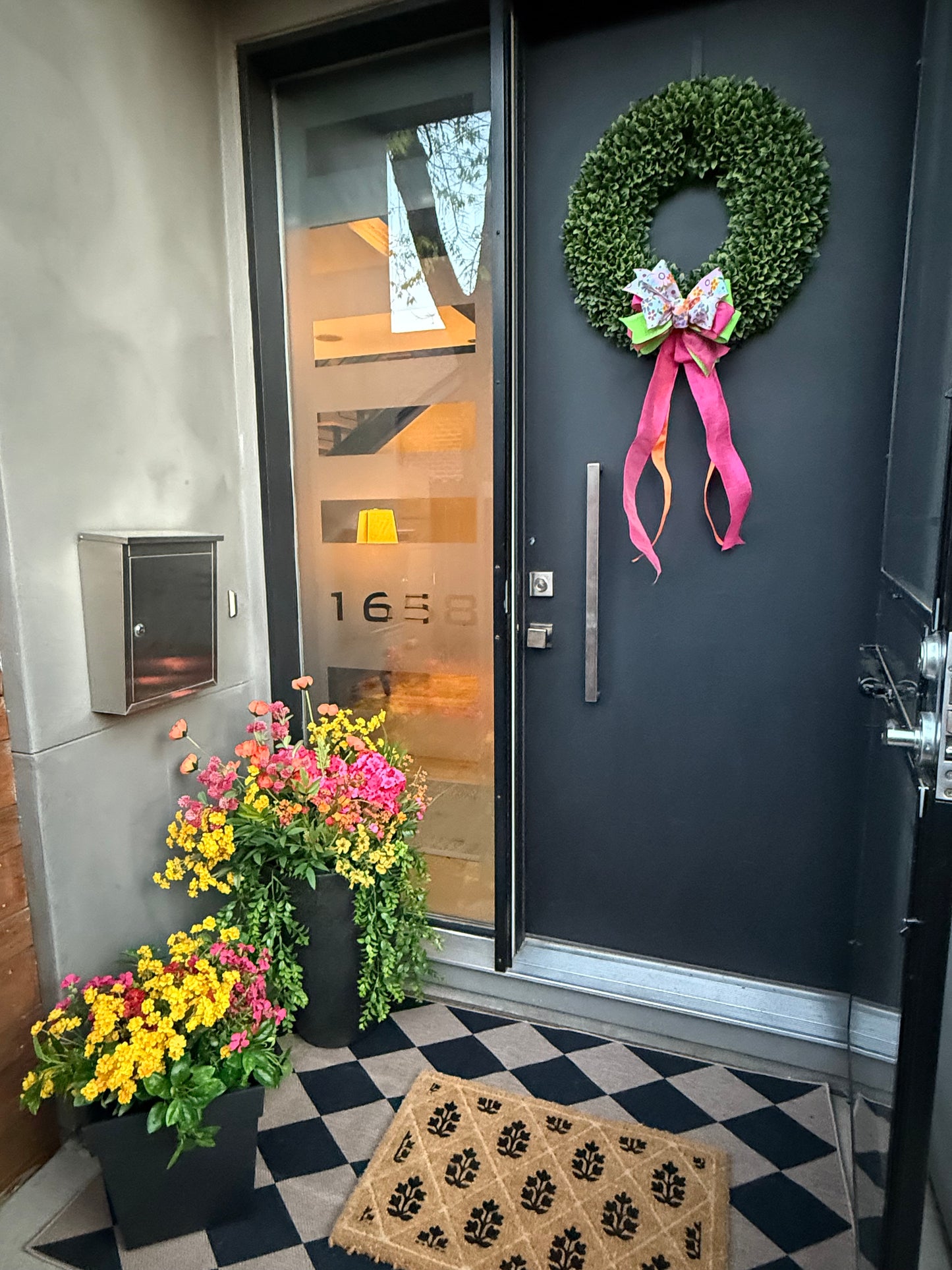 Decorative wreath with a pink ribbon on a door, flowers on the ground, and a checkered rug.
