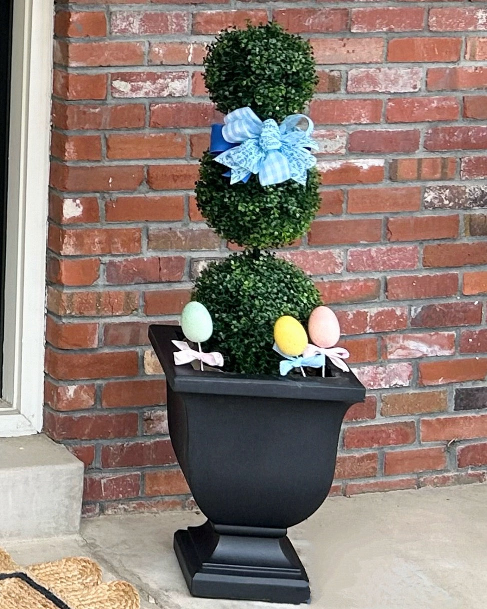 A boxwood topiary in a black pedestal planter, decorated with a blue bow and Easter eggs.