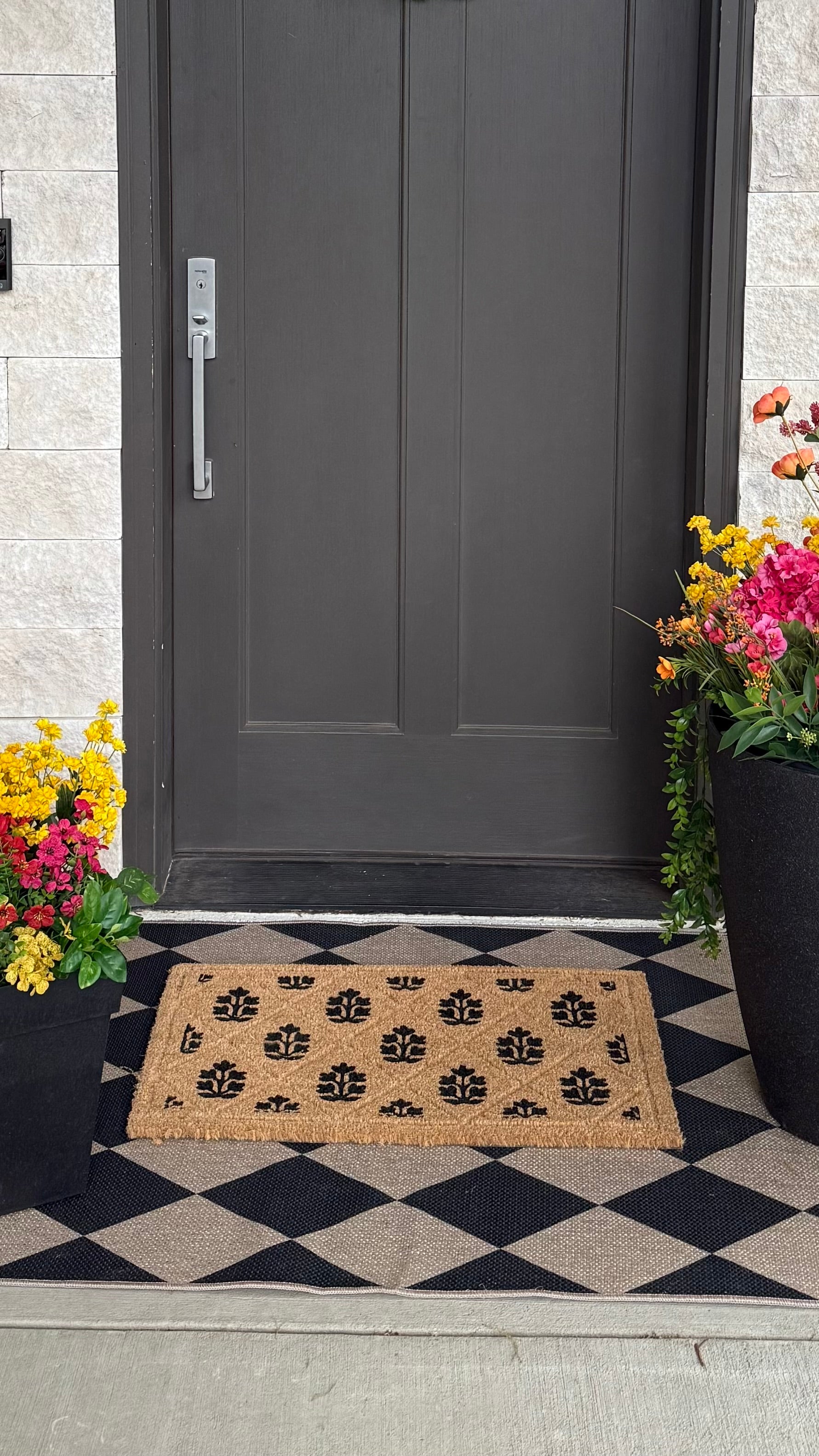 Gray door with a doormat, flowers, and a decorative rug on a checkered floor.