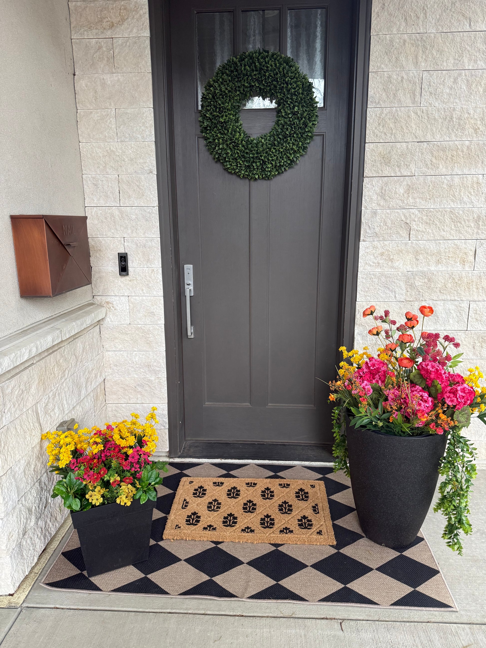 Front door with a wreath, potted plants, and a doormat on a checkered rug.