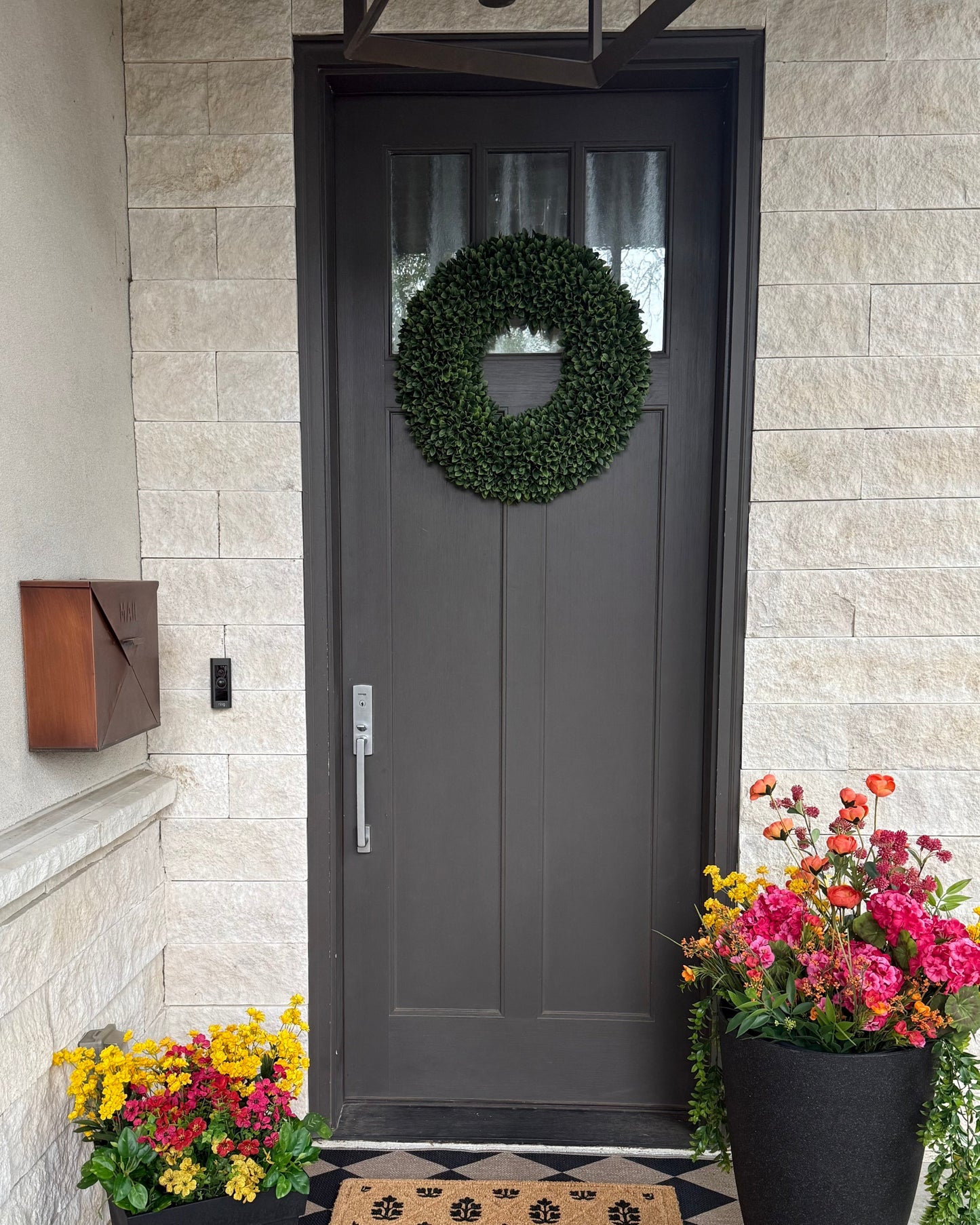 Front door of a house with decorative wreath, flowers, and doormat.