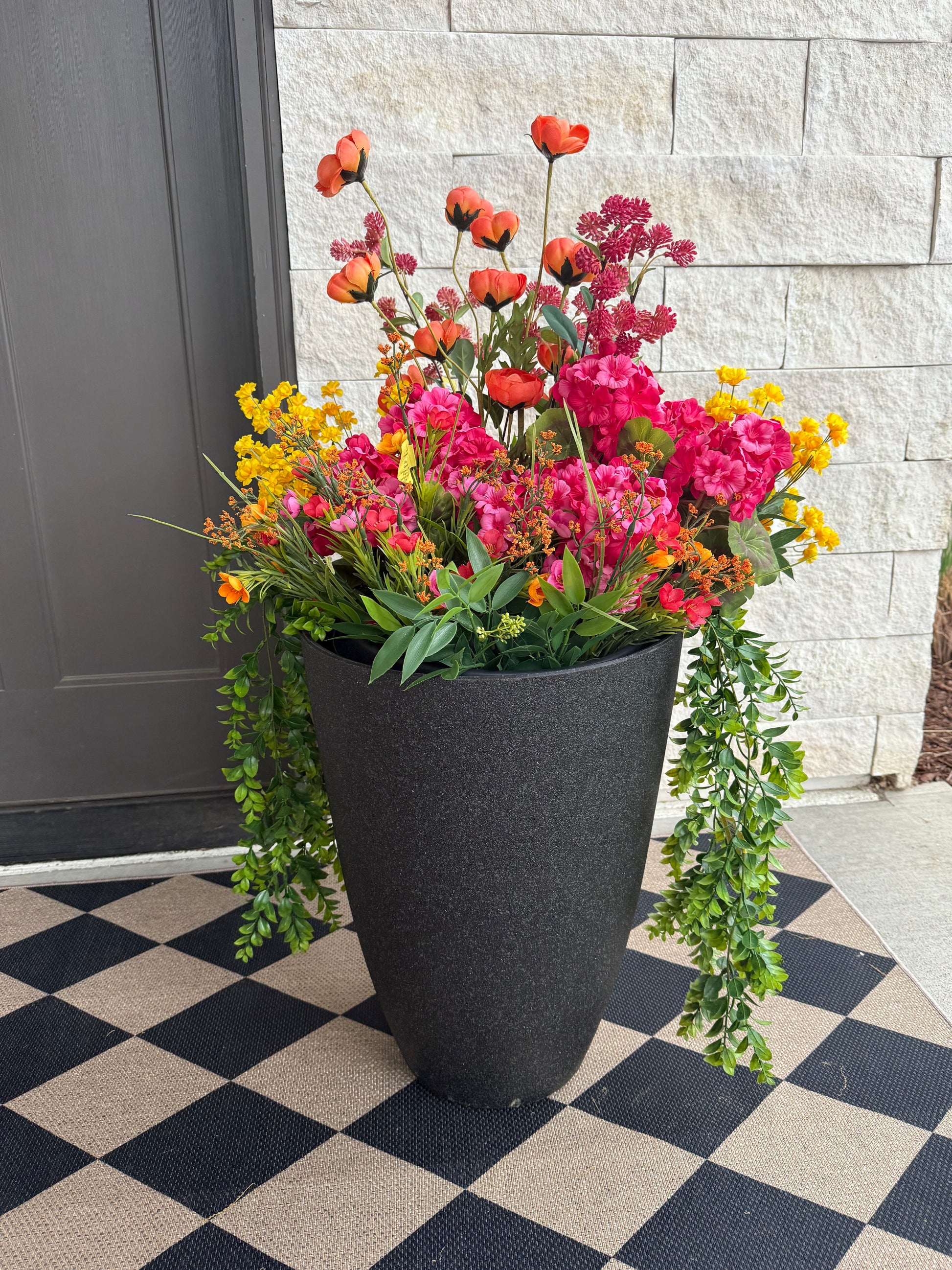 A tall black resin flower planter burst with vibrant artificial faux florals, placed on a checkered black and white floor mat, against a stone wall background.