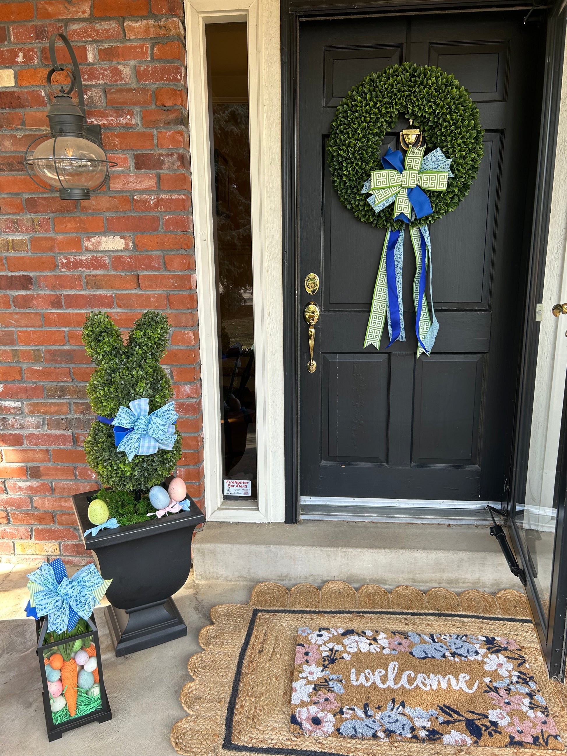 A front porch decorated for Spring with a green boxwood wreath, blue and white bow, a boxwood bunny topiary in a black planter, a 'Welcome' doormat, and a hurricane lantern.