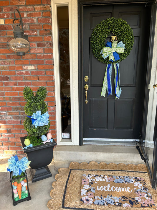 A front porch decorated for Spring with a green boxwood wreath, blue and white bow, a boxwood bunny topiary in a black planter, a 'Welcome' doormat, and a hurricane lantern.